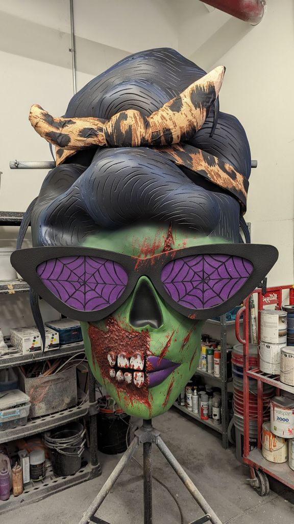 Completed custom female zombie head with beehive hairdo, purple spider web glasses, purple lips, blood splatter, and torn cheek with exposed teeth