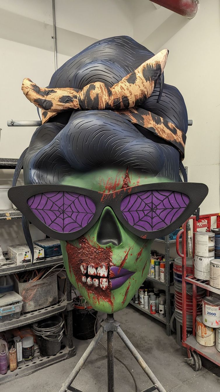 Completed custom female zombie head with beehive hairdo, purple spider web glasses, purple lips, blood splatter, and torn cheek with exposed teeth