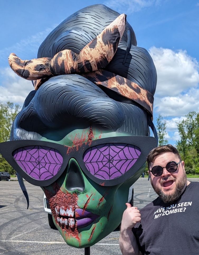 Selfie opportunity with custom female zombie head with beehive hairdo, purple spider web glasses, purple lips, blood splatter, and torn cheek with exposed teeth